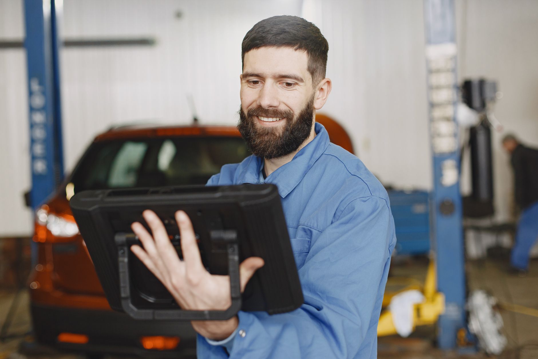 A smiling mechanic in a blue uniform using a tablet for vehicle diagnostics