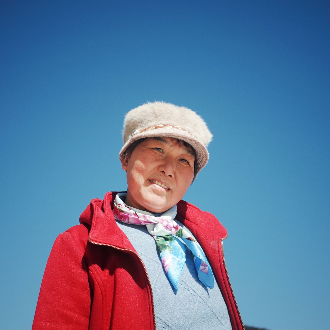 A woman in a red jacket and beige hat smiling under a blue sky