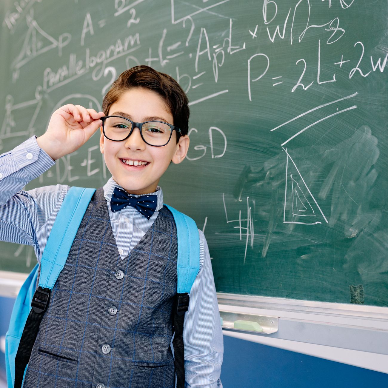 A smiling boy in a bow tie and backpack by a blackboard