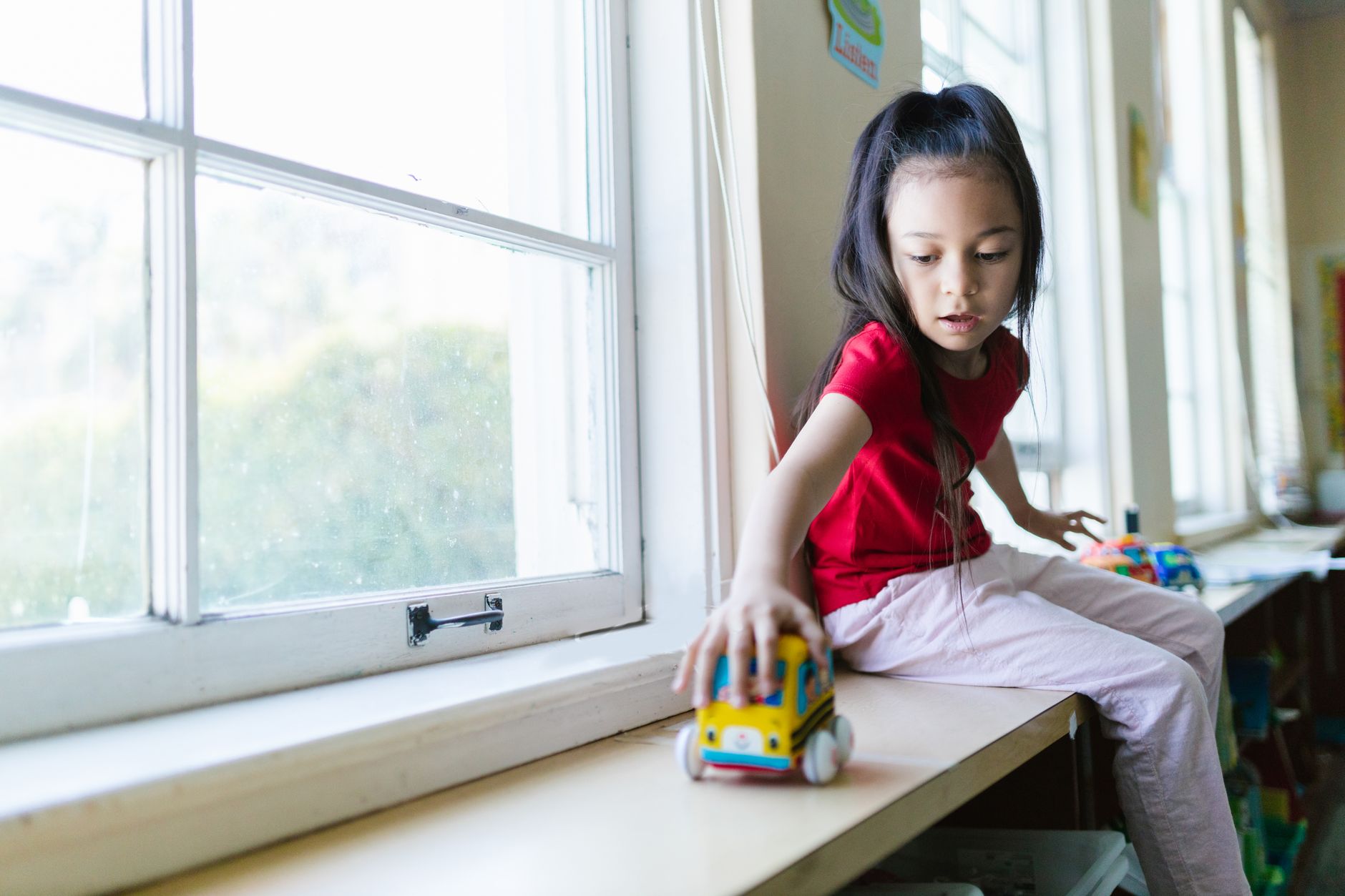 A young girl in a red shirt plays with a toy beside a window in a sunny classroom setting.
